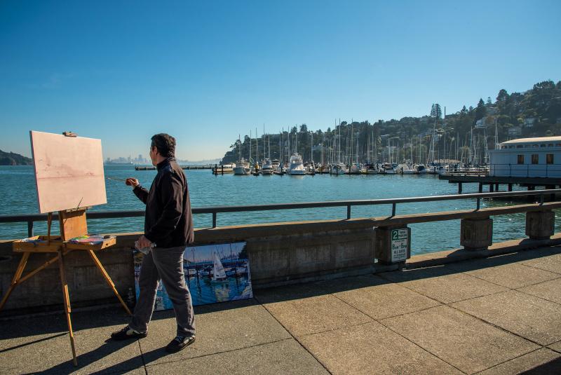 A person painting near the Bay with boats in the background